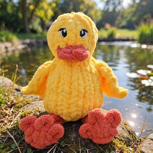 May include: A handmade crochet plush duck toy featuring a bright yellow body with a textured, soft yarn finish. The duck has a prominent orange beak and matching orange webbed feet. It includes embroidered black eyes with white highlights.