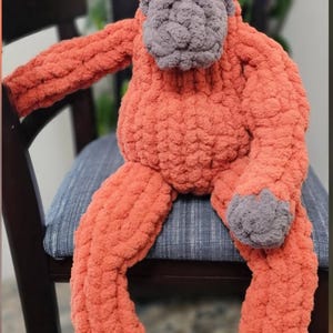 May include: A handmade crochet orangutan plush toy crafted from thick, textured orange yarn. The figure features a prominent gray face, gray hands, and gray feet, with dark eyes set into the face. The plush has long arms and legs.