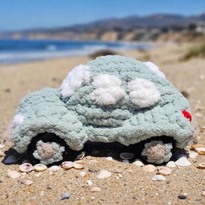 Könnte beinhalten: Ein gehäkeltes Plüsch-Spielzeugauto in sanftem Salbeigrün mit weißen, flauschigen, wolkenartigen Akzenten auf dem Dach und den Seiten. Die Räder sind dunkelbraun mit strukturierten Mitten und ein kleines rotes Detail ist am Heck des Fahrzeugs sichtbar.