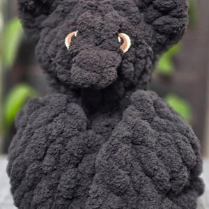 May include: A plush toy shaped like a bat, crafted from thick, textured black yarn. The creature features large, rounded ears and small, almond-shaped eyes with white and brown accents. The body is rounded and covered in a soft, fuzzy material.