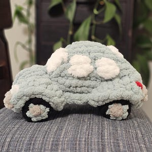 Könnte beinhalten: Ein gehäkeltes Plüschauto-Spielzeug in sanftem Salbeigrün mit weißen, strukturierten Wolken auf dem Dach und an den Seiten. Das Fahrzeug hat schwarze Räder mit beigen Mitten und einen kleinen roten Akzent am Heck. Das Garn hat eine dicke Textur.
