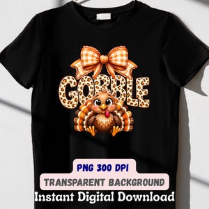 May include: A black t-shirt featuring a graphic of a cartoon turkey with brown feathers, a red wattle, and large eyes. Above the turkey is the word GOBBLE in a leopard print pattern, topped with a large orange and white checkered bow.
