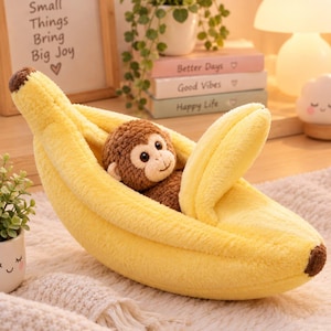 May include: A plush yellow banana-shaped bed containing a small brown and cream-colored stuffed monkey toy. The banana has a soft, fuzzy texture with a brown tip at each end and a curved, open peel design that cradles the plush monkey inside.