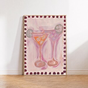 Op de afbeelding: Een ingelijst schilderij met twee martiniglazen gevuld met oranje en paarse vloeistof, gegarneerd met schijfjes citrusvrucht. Het kunstwerk is geplaatst tegen een lichtroze achtergrond en omrand door een decoratieve rand van donkerrode markeringen.