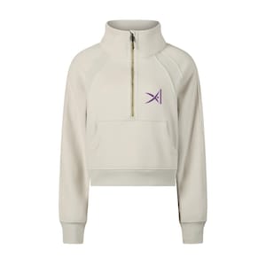 Könnte beinhalten: Ein beiges, bauchfreies Sweatshirt mit hohem Kragen und goldenem Halb-Reißverschluss. Das Kleidungsstück verfügt über lange Ärmel mit gerippten Bündchen, eine Kängurutasche auf der Vorderseite und ein violettes, abstraktes Logo auf der linken Brust.