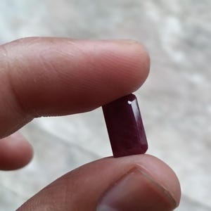 May include: A deep red, rectangular-cut gemstone with faceted edges. The stone has a translucent quality and a polished surface, displaying a rich, saturated crimson hue throughout its structure, held between two fingers for display.