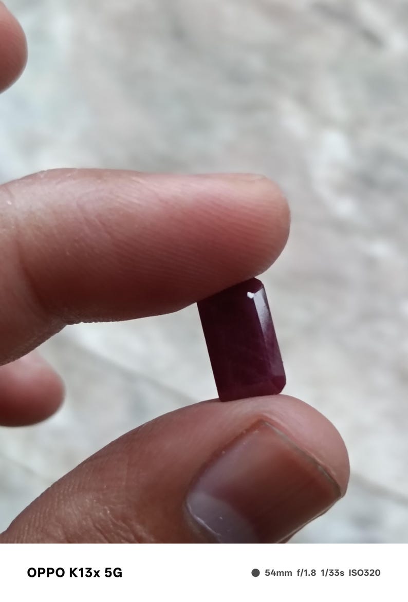 May include: A deep red, rectangular-cut gemstone with faceted edges. The stone has a translucent quality and a polished surface, displaying a rich, saturated crimson hue throughout its structure, held between two fingers for display.