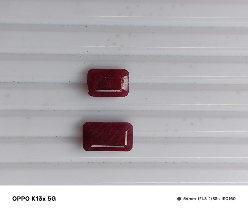 May include: Two rectangular, deep red gemstones with faceted surfaces. The stones are positioned one above the other, displaying a rich, saturated crimson hue and a polished finish. The text OPPO K13x 5G and camera settings are visible at the bottom.