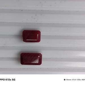 May include: Two rectangular, deep red gemstones with faceted surfaces. The stones are positioned one above the other, displaying a rich, saturated crimson hue and a polished finish that reflects light across their geometric, multi-faceted edges.