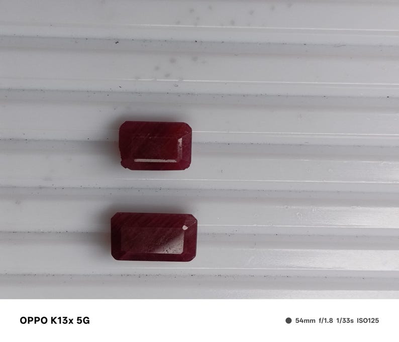 May include: Two rectangular, deep red gemstones with faceted surfaces. The stones are positioned one above the other, displaying a rich, saturated crimson hue and a polished finish that reflects light across their geometric, multi-faceted edges.
