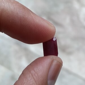 May include: A small, rectangular, deep red gemstone with faceted edges. The stone has a polished surface and a rich, saturated crimson color. It is held between two fingers, showcasing its compact size and geometric cut against a neutral background.
