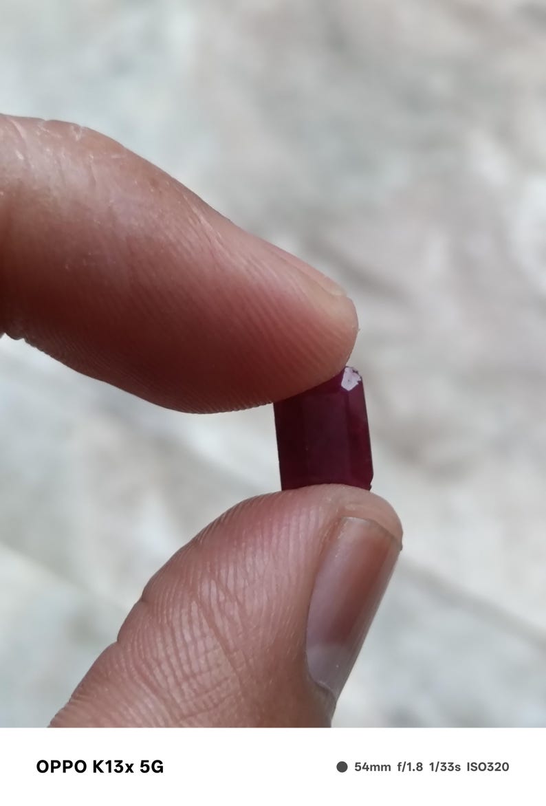 May include: A small, rectangular, deep red gemstone with faceted edges. The stone has a polished surface and a rich, saturated crimson color. It is held between two fingers, showcasing its compact size and geometric cut against a neutral background.