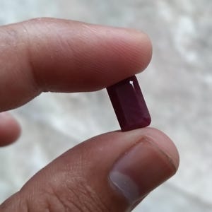 May include: A deep red, rectangular gemstone with faceted edges. The stone has a smooth, polished surface that reflects light, showcasing a rich, saturated crimson hue throughout its elongated, geometric shape and clean, sharp corners.