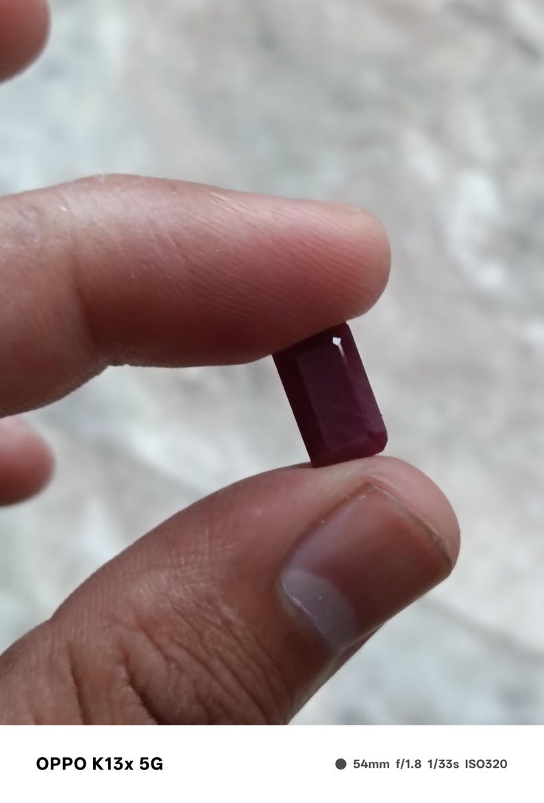 May include: A deep red, rectangular gemstone with faceted edges. The stone has a smooth, polished surface that reflects light, showcasing a rich, saturated crimson hue throughout its elongated, geometric shape and clean, sharp corners.
