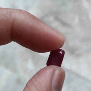 May include: A small, rectangular, faceted gemstone with a deep, saturated red color. The stone features sharp, clean edges and a polished surface that reflects light, showcasing its translucent quality and precise geometric cut.