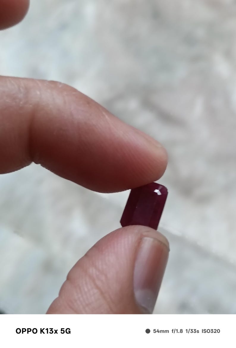 May include: A small, rectangular, faceted gemstone with a deep, saturated red color. The stone features sharp, clean edges and a polished surface that reflects light, showcasing its translucent quality and precise geometric cut.