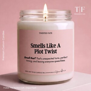 May include: A clear glass jar candle with a lit wick and white wax. The label is light pink with black text reading Twisted Fate, Smells Like A Plot Twist, and 100% Soy Wax Candle 9oz. The label also includes descriptive text about unexpected turns.