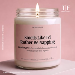 May include: A 9oz soy wax candle in a clear glass jar with a lit wick. The pink label features the text: Smells Like I'd Rather Be Napping. Smell that? That's canceled plans, cozy blankets, and absolutely zero regrets. Twisted Fate branding.