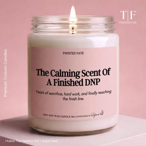 May include: A 9oz soy wax candle in a clear glass jar with a lit wick. The pink label features the text: TWISTED FATE, The Calming Scent Of A Finished DNP, Years of sacrifice, hard work, and finally reaching the finish line, 100% SOY WAX CANDLE 9oz.