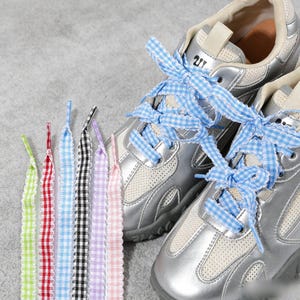 Puede incluir: Un par de zapatillas deportivas plateadas metalizadas con paneles de malla y cordones de cinta de cuadros vichy azul claro, acompañadas de un juego de seis cordones de cinta planos adicionales en cuadros vichy verde lima, rojo, azul, negro, lavanda y rosa.