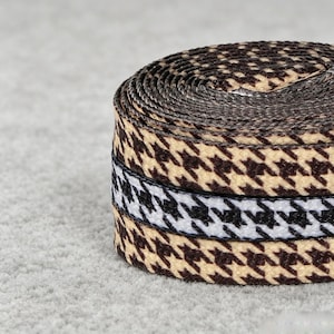 May include: A coiled fabric ribbon featuring a repeating houndstooth pattern. The design alternates between sections of dark brown and beige houndstooth and a central band of black and white houndstooth, creating a textured, woven appearance.