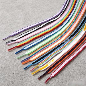 May include: A collection of flat shoelaces featuring a variety of colors and patterns, including solid purple, black and white, pink, rainbow stripes, blue, green, orange, and red, each finished with a plastic aglet at the end of the lace.