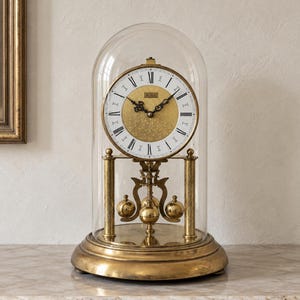 Könnte beinhalten: Eine goldfarbene Jahresuhr mit einem runden weißen Zifferblatt, schwarzen römischen Ziffern und einer strukturierten goldenen Mitte. Die Uhr befindet sich unter einer klaren Glaskuppel und steht auf einem runden Messingsockel mit Drehpendel.