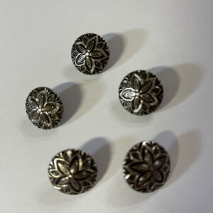 May include: Five silver-colored buttons with a floral design. The buttons are round with a detailed, embossed pattern of petals and leaves. The buttons are arranged on a white surface.
