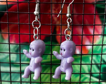 May include: A pair of drop earrings featuring small, light purple plastic baby doll charms. Each charm is suspended from a silver-toned metal fishhook wire by a series of small circular jump rings. The dolls have painted black eyes and red lips.