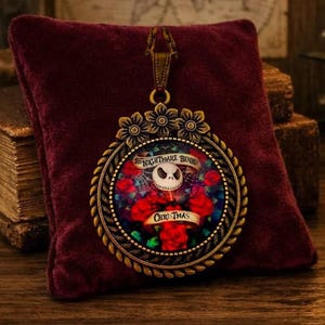 Może przedstawiać: Okrągły wisiorek w kolorze brązu z centralnym obrazem czaszki otoczonej czerwonymi różami i pajęczynami. Tekst "The Nightmare Before Christmas" jest nadrukowany na banerach na obrazie. Ramka ma krawędź przypominającą skręconą linę.