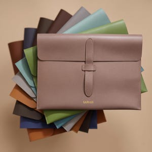 May include: A mauve document folder with a vertical strap closure and the name SARAH embossed in gold. The folder is displayed on top of a stack of similar folders in various colors including brown, blue, green, and gray, arranged in a fan shape.