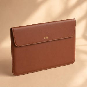 May include: A rectangular brown leather laptop sleeve with a fold-over flap closure. The center of the flap features the gold embossed letters CM. The sleeve has a smooth, matte finish with clean, stitched edges along the entire perimeter.