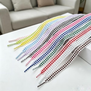 May include: A collection of flat shoelaces featuring a gingham check pattern in various colors including yellow, light blue, lavender, pink, dark blue, green, red, grey, and brown, each finished with a white scalloped edge trim along both sides.