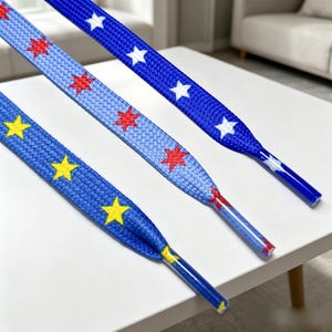 May include: Three flat shoelaces with aglets. One lace is dark blue with white stars, one is light blue with red stars, and one is medium blue with yellow stars. The laces feature a woven texture and are arranged diagonally across the frame.