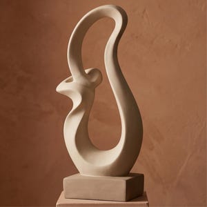 Può includere: Una scultura in ceramica beige caratterizzata da una forma verticale alta, fluida e astratta con un'apertura centrale. La scultura è montata su una solida base rettangolare e presenta una finitura liscia e opaca con linee curve.