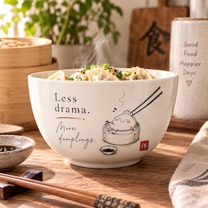 Może przedstawiać: Biała ceramiczna miska wypełniona pierożkami na parze, z czarnym nadrukiem "Less drama. More dumplings." oraz grafiką uśmiechniętego pierożka w bambusowym koszyku, podnoszonego pałeczkami, z małą czerwoną pieczątką w kształcie kwadratu.