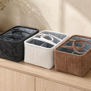 May include: Three rectangular storage baskets made of corduroy fabric in charcoal gray, white, and brown. Each basket features a central divider, two open compartments, and two fabric handles attached to the top rim, with a striped interior lining.