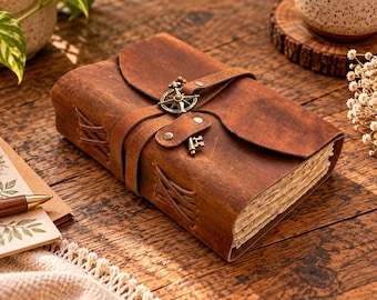 May include: A brown leather journal featuring a wrap-around strap secured with a metal compass charm and a small key accent. The journal has visible side stitching, a textured cover, and thick, deckled-edge pages, presenting a classic, durable design.