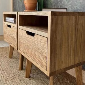 May include: Two light brown wooden bedside tables with a visible grain pattern, each featuring a single drawer with a recessed handle and an open storage shelf above. The bedside tables are supported by four tapered wooden legs.