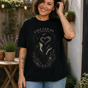 Puede incluir: Una camiseta negra de manga corta con un estampado gráfico de una persona con pelo largo rodeada de elementos florales, estrellas y una forma de corazón. El texto "LOVE CAN DO EVERYTHING" está impreso en arco sobre la ilustración.