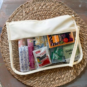 May include: A clear rectangular vinyl pouch with a white zipper and wrist strap containing various personal care items including a Shout stain remover wipe, a pink Ultima electrolyte packet, a pack of Kleenex tissues, and small green Tic Tac mints.