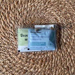 May include: A clear plastic pouch with a metal snap closure and a ball chain, containing a Brookstone makeup remover towelette with hyaluronic acid and eucalyptus, measuring 7.2 inches by 6.4 inches, alongside a deodorant wipe packet.
