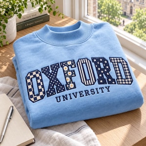 Puede incluir: Una sudadera azul claro de cuello redondo con la palabra OXFORD en letras grandes de estilo bloque. Cada letra tiene un patrón único, incluyendo cuadros, lunares, margaritas, rayas y estrellas. La palabra UNIVERSITY está bordada debajo.