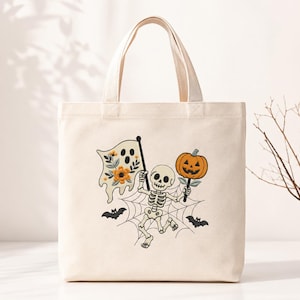 Op de afbeelding: Een crèmekleurige canvas tas met een centrale afbeelding van een skelet dat op een spinnenweb staat. Het skelet houdt in de ene hand een vlag met een geest en bloemen vast en in de andere een oranje pompoen, vergezeld door twee kleine zwarte vleermuizen.