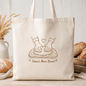 Puede incluir: Un bolso de tela color crema con una ilustración de líneas marrones de dos manos amasando masa sobre una tabla de madera, un pequeño corazón y el texto "Knead More Bread" con espigas de trigo decorativas a los lados.