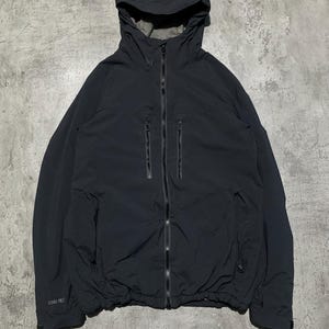 May include: A charcoal black hooded jacket featuring a full-length front zipper, two vertical zippered chest pockets, and an adjustable drawstring hem. The sleeve cuff displays a small Gore-Tex logo, and the interior lining is a contrasting grey.