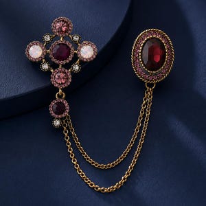 May include: A gold-toned brooch featuring a cross-shaped component with deep red, pink, and iridescent crystals, connected by two delicate gold chains to a separate oval-shaped piece centered with a large, dark red faceted gemstone.