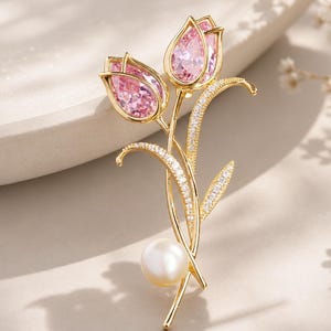 May include: A gold-toned brooch shaped like two tulip flowers with faceted pink crystal petals. The stems and leaves are accented with small clear pavé stones, and a single round white pearl is positioned at the base of the stems.