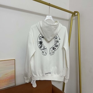 May include: A white hooded sweatshirt featuring a large black horseshoe-shaped graphic on the back with intricate gothic-style lettering and decorative details. The hoodie includes a drawstring hood, long sleeves, and a small logo near the hem.