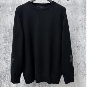 May include: A solid black long-sleeve sweater featuring a ribbed crew neckline, ribbed cuffs, and a ribbed hem. Each sleeve is adorned with a dark, cross-shaped leather patch positioned at the elbow area, providing a distinct textural contrast.